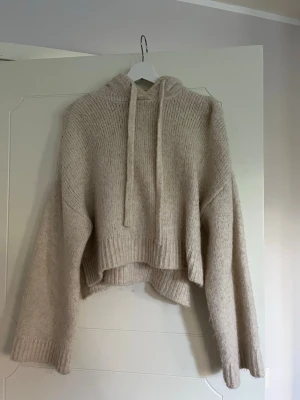Beige stickad hoodie med luva - Mysig beige stickad hoodie med luva och dragsko. Tröjan har en croppad passform, breda ärmar och ribbade muddar vid ärmslut och nederkant. Perfekt för lager på lager och chill dagar. Materialet känns mjukt och fluffigt.