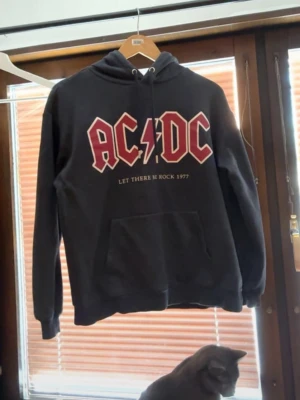 Svart AC/DC hoodie med tryck - Svart hoodie med stor AC/DC-logga i rött och vitt på bröstet samt texten 'Let There Be Rock 1977' under. Klassisk känguruficka framtill och justerbar huva med snören. Perfekt för dig som gillar rock och vill ha en bekväm och snygg tröja.
