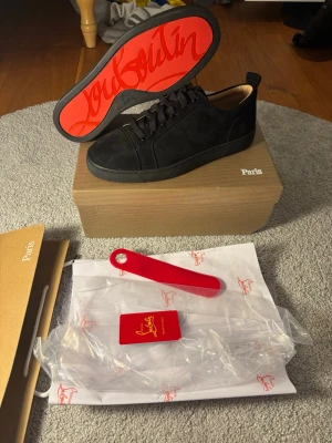 Svarta Christian Louboutin sneakers - Snygga svarta sneakers från Christian Louboutin med klassisk röd sula och diskret logga. Skorna har lågt skaft, ovandel i mocka och svarta skosnören. Perfekta för dig som vill sticka ut med exklusiv design och ikoniska detaljer.