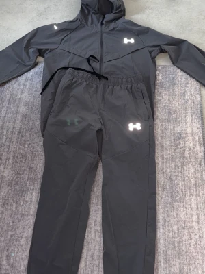 Svart träningsset från Under Armour - Snyggt svart träningsset från Under Armour med matchande jacka och byxor. Jackan har huva och dragkedja, medan byxorna har resår i midjan. Båda plaggen har reflekterande loggor och är tillverkade i ett lätt, tekniskt material som passar perfekt för träning.