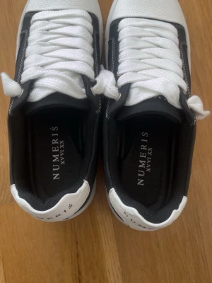 Svarta och vita sneakers från Numeris - Snygga sneakers från Numeris i svart och vitt med klassisk låg profil. Skorna har vita snören, svart innerfoder och detaljer i både svart och vitt. Tillverkade i Portugal och har en stilren design som passar till många olika outfits.
