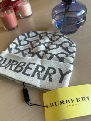 Vit och grå Burberry mössa - Snygg vit mössa från Burberry med grått logomönster och stor Burberry-text på uppvikt kant. Klassisk beanie-modell i mjukt material, perfekt för att lyfta din outfit med en exklusiv touch.