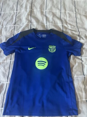 Blå FC Barcelona träningströja Nike - Snygg blå FC Barcelona träningströja från Nike med neongula detaljer, klubbmärke och sponsorlogga på bröstet. Tröjan har korta ärmar och är tillverkad i ett lätt och ventilerande material, perfekt för träning eller match.