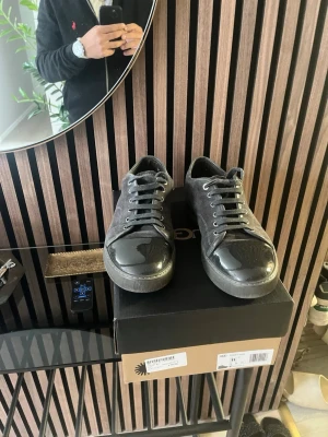 Svarta sneakers från Lanvin - Stilrena svarta sneakers från Lanvin med mockaliknande material på sidorna och glansig tå i läder. Klassisk låg modell med svarta skosnören och diskreta sömmar. Perfekta för dig som gillar en clean och modern look.