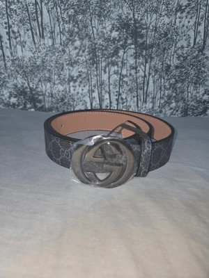 Svart Gucci bälte med GG-spänne - Snyggt svart bälte från Gucci med klassiskt GG-mönster och stort metallspänne med GG-logga. Bältet har en stilren design och insidan är beige. Perfekt accessoar för att lyfta vilken outfit som helst.