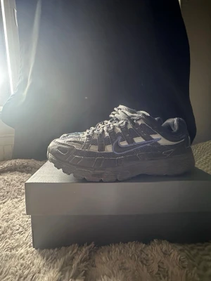 Nike p-6000svart/grå/blå - Nike P-6000sneakers i svart och grått med meshpaneler och detaljer i syntet. Skon har en chunky sula och klassisk Nike Swoosh på sidan. Skön passform med vadderad plös och häl, samt snörning framtill. Perfekt för dig som gillar sportig och trendig stil.