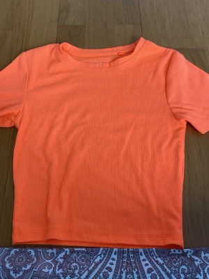 Ribbad orange t-shirt - En ribbad t-shirt i en stark orange färg. T-shirten har korta ärmar och rund halsringning. Materialet känns mjukt och stretchigt, perfekt för en avslappnad stil. Enkel och clean design utan tryck eller logga.