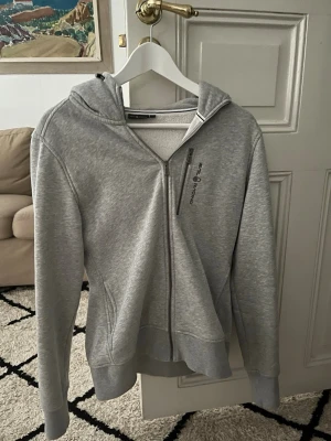 Grå hoodie från Sail Racing - Snygg grå hoodie från Sail Racing med dragkedja framtill och huva. Tillverkad i mjukt material med borstad insida för extra komfort. Samt att den passar på nån med Small i storlek med tanken på att det är L i barnstorlek!!