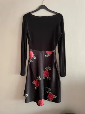 Svart klänning med rosor  - Svart klänning från Hennes Collection med långa ärmar och har ett blommigt mönster med röda rosor och gröna blad. Klänningen är i storlek S och är i väldigt bra skick.