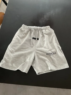 Grå Essentials Fear of God shorts - Snygga ljusgrå shorts från Essentials Fear of God med elastisk midja och snörning. Mjuk bomullskänsla och diskret logga på vänster ben. Perfekta för chill dagar eller streetwear-stil.