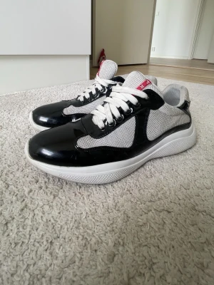 Svart Pra”da cups  - Snygga sneakers från Pra”da i svart och vitt med glansiga svarta detaljer och meshpaneler. Skorna är i acceptabelt skick. Höger skon bakdel är lite sämre skick än den vänstra skon men kan fixas. Snören är också lite smutsig men kan fixas i tvätten.