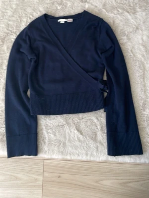 Mörkblå omlottstickad tröja - Snygg mörkblå stickad omlottopp med långa ärmar och bred ribbad mudd i nederkant och vid ärmslut. Tröjan knyts i sidan för en trendig look och har en djup v-ringning. Perfekt att styla med jeans eller kjol. Använt max 2 gånger 