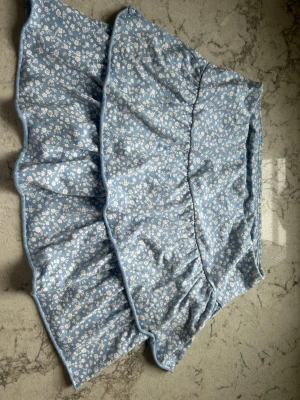 Blå blommig volangkjol från Lindex - Supersöt blå kjol från Lindex med vitt blommönster och volangdetaljer. Kjolen har en kort modell och är gjord i mjuk bomull, perfekt för en fräsch och somrig look.