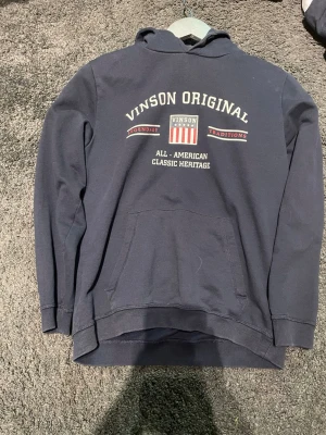 Mörkblå hoodie från Vinson Original - Mörkblå hoodie från Vinson Original med tryck på bröstet i vitt och rött. Klassisk känguruficka framtill och huva. Snygg streetwear-stil med texten 'All-American Classic Heritage' och ribbade muddar vid ärmslut och nederkant.