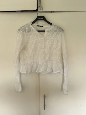 Vit smockad blus från 157 - Supersöt vit blus från 157 med smockdetaljer över hela och volangkant nedtill. Blusen har knappar framtill och långa ärmar. Perfekt till jeans eller kjol för en fräsch look.