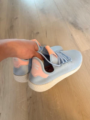 Ljusblå Adidas sneakers med rosa detaljer - Säljer ett par snygga ljusblå Adidas sneakers med rosa detaljer på häl och plös. Skorna har en vit, strukturerad sula och är tillverkade i meshmaterial som andas. Perfekta för dig som vill ha en fräsch och sportig look. Klassisk låg modell med snörning.