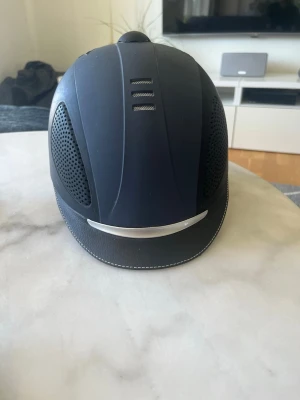 Svart ridhjälm med ventilationsnät - Snygg svart ridhjälm med ventilerande nät på sidorna och en silverfärgad detalj framtill. Hjälmen har en matt finish och stilrena sömmar längs skärmen. Perfekt för dig som vill ha både säkerhet och stil i stallet.