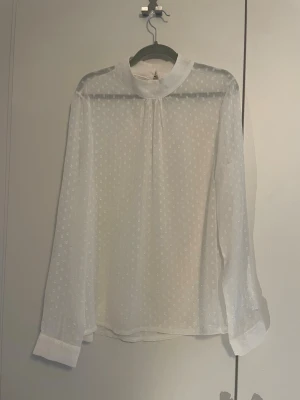 Vit blus - Säljer en vit, transparent blus med diskret prickigt mönster och hög krage. Blusen har långa ärmar med breda manschetter och knäppning i nacken. Tillverkad i ett lätt och luftigt material som ger en elegant look.