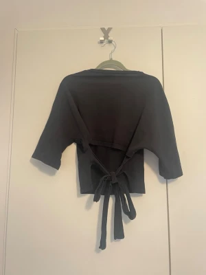 Knyt t-shirt - Knyt topp från Nelly. Snygg svart omlottopp med trekvartsärm och långa knytband i midjan som kan knytas bak eller fram. Toppen har en enkel, stilren design och är gjord i mjukt material som känns skönt mot huden. Perfekt för dig som gillar trendiga och bekväma plagg.