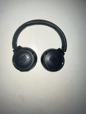 Svarta on-ear hörlurar från JBL - Snygga svarta on-ear hörlurar från JBL med vadderade öronkuddar och justerbart huvudband. Hörlurarna har en stilren design i plast och konstläder, perfekt för musik och gaming. JBL-loggan syns tydligt på båda sidorna.