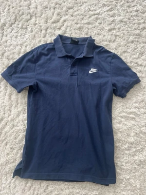 Marinblå pikétröja från Nike XS - Snygg marinblå pikétröja från Nike i storlek XS. Klassisk krage, korta ärmar och vit Nike-logga broderad på bröstet. Tillverkad i mjuk bomull som känns skön mot huden. Perfekt för en sportig och avslappnad stil.