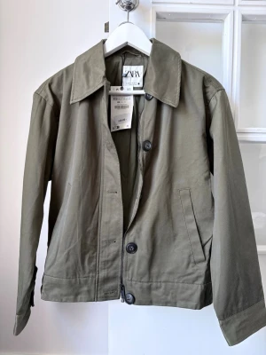 Olivgrön jacka från Zara - Snygg olivgrön jacka från Zara i storlek XS med klassisk krage och raka linjer. Jackan har knappar framtill, två sidofickor och är tillverkad i bomull. Perfekt för dig som gillar minimalistisk stil och vill ha en enkel men trendig jacka. Aldrig använd med prislapp på! Går att både knäppa med kanppar samt dragkedja och tillkommer en extra knapp.