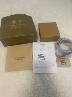 Burberry beige rutigt bälte med guldspänne - Snyggt Burberry-bälte i klassiskt beige och rutigt mönster med ett stort guldfärgat 'B'-spänne. Bältet kommer med originalpåse, box och kvitto. Perfekt accessoar för att lyfta din outfit med en lyxig touch.