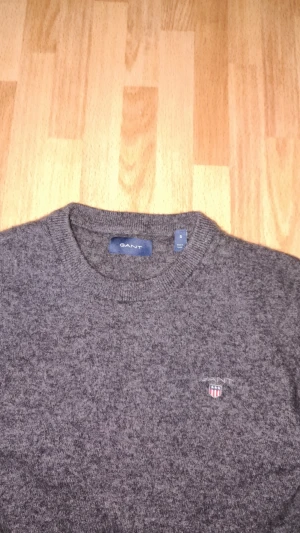 Mörkgrå stickad tröja från GANT - Mörkgrå stickad tröja från GANT med rund halsringning och diskret broderad logotyp på bröstet. Tröjan har långa ärmar och är tillverkad i mjukt material som passar perfekt för lager på lager. Klassisk och stilren design.