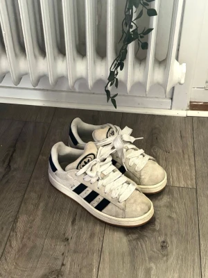Adidas campus 00 - Klassiska vita Adidas sneakers med svarta ränder på sidorna och Adidas-logga på plösen. Skorna har en låg profil, vita skosnören och yttersula i gummi. Tillverkade i mocka och textil för en stilren och sportig look.