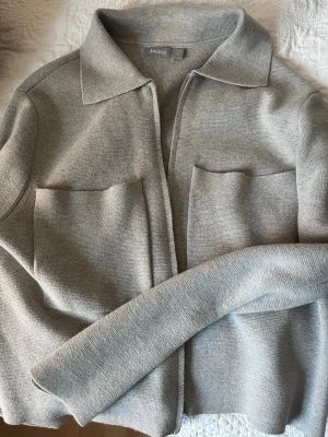 Ljusgrå cardiganjacka från Åhléns - Snygg ljusgrå cardiganjacka från Åhléns i stickad kvalitet. Jackan har två stora framfickor, krage och dragkedja framtill. Enkel att matcha med olika outfits. Jackan säljs på Åhléns för 599:-