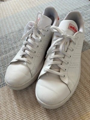 Vita Adidas sneakers med röd detalj - Snygga vita sneakers från Adidas med klassisk design och röda detaljer på häl och plös. Skorna har perforerade sidor för ventilation och vita skosnören. Mjukt innerfoder och cloudfoam comfort-sula för extra komfort. Perfekta för en clean och sportig look.