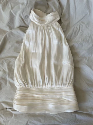 Vit halterneck topp i satin - Har en liten reva på ena sidan men inget som syns!! Snygg vit halterneck topp i glansigt satinliknande material med bred plisserad kant nedtill och hög krage. Toppen har en elegant, lös passform och knäpps i nacken med knapp. Perfekt för dig som vill sticka ut med en stilren look. 