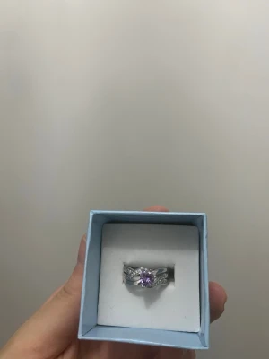 Ring i silverfärg med lila sten - Snygg silverfärgad ring med ett lila hjärta i mitten och små klara kristaller längs bandet. Ringen har en tvinnad design som ger ett elegant intryck. Färgen ska hålla om den inte kommer i kontakt med vatten, parfym/parfymerade krämer och tror alkogel?