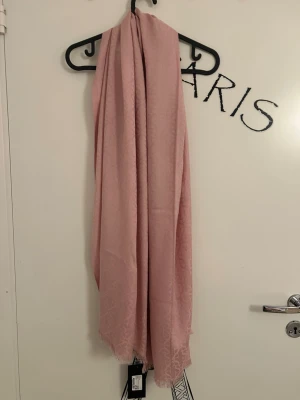 Ljusrosa scarf från Stockh LM - Ljusrosa scarf med diskret mönster och fransad kant från Stockh LM. Scarfen är rektangulär och har ett mjukt tyg som är i 100% viskos. Helt oanvänd med prislappen kvar. Nypris på scarfen är 499 kr.  