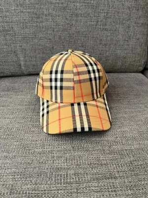 Beige rutig keps från Burberry - Helt oanvänd. Säljer en klassisk beige keps från Burberry med det ikoniska rutmönstret i svart, vitt och rött. Kepsen har böjd skärm och justerbar rem bak. Tillverkad i bomull för en bekväm och snygg look. Perfekt accessoar för dig som gillar streetstyle och lyxig känsla.