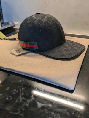 Svart keps med GG-mönster och rand - Snygg svart keps från Gucsi med klassiskt GG-monogram över hela kepsen. På sidan finns en grön och röd rand som ger en ikonisk touch. Justerbar rem baktill för perfekt passform. Materialet är tyg med exklusiv känsla.