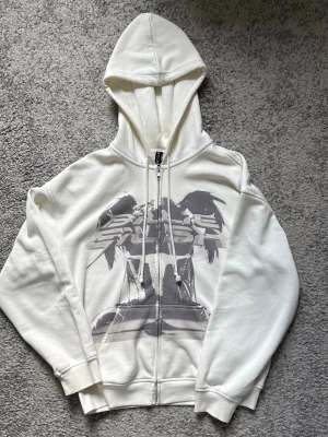 Vit Billie Eilish zip hoodie - Säljer en vit zip hoodie från Billie Eilish med tryck framtill i grått och svart. Hoodien har huva med dragsko, långa ärmar och ribbade muddar. Perfekt för dig som gillar streetwear och vill ha något unikt från 2023.