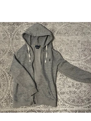 Grå hoodie från Polo Ralph Lauren - Snygg grå hoodie från Polo Ralph Lauren med dragkedja, vita dragsnören och klassisk logga broderad på bröstet. Tillverkad i mjukt sweatshirtmaterial och har två fickor framtill samt ribbade muddar vid ärmar och nederkant. Perfekt för en chill och stilren look.