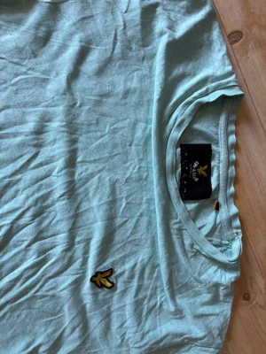Ljusblå t-shirt från Lyle & Scott - Snygg ljusblå t-shirt från Lyle & Scott med klassisk gul logga på bröstet. T-shirten har rund halsringning och korta ärmar. Perfekt till jeans eller shorts för en avslappnad stil.