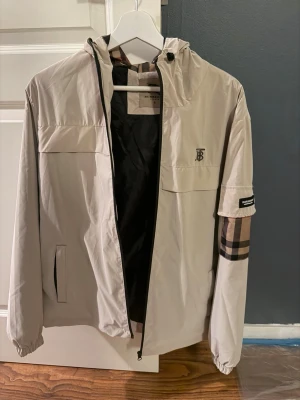 Beige vindjacka från Burberry - Stilren beige vindjacka från Burberry med klassiskt rutigt mönster på ena ärmen och svart innerfoder. Jackan har huva med dragsko, dragkedja framtill och elastiska muddar. Burberry-logga på bröstet och patch på ärmen ger en exklusiv känsla.