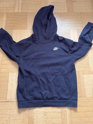 Svart Nike hoodie med logga - Svart hoodie från Nike med klassisk vit logga på bröstet. Hoodien har huva och en stor magficka framtill. Tillverkad i mjukt material som passar perfekt för en avslappnad stil. Ribbade muddar vid ärmslut och nederkant.