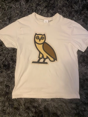 Beige OVO t-shirt - Säljer en beige t-shirt med ett stort tryck av en stiliserad uggla i brunt och gult på framsidan. T-shirten har rund hals och korta ärmar. Perfekt för dig som gillar unika prints och enkel stil.Köpt den från kanada själva affären i vancover har liten fläck som man ser i sista bilden den är i size S men passar mer M och L.  För frågor är det vara skriva☺️