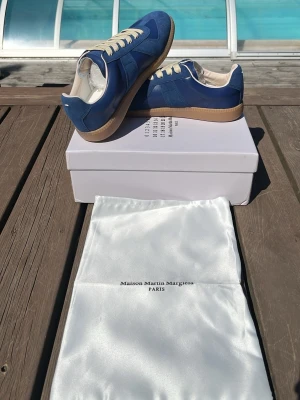 Blå Maison Margiela sneakers - Snygga blå sneakers från Maison Martin Margiela med beige snörning och gummisula. Skorna har en klassisk låg siluett och är tillverkade i en mix av mocka och läder. Perfekta för dig som vill ha en clean och stilren look.