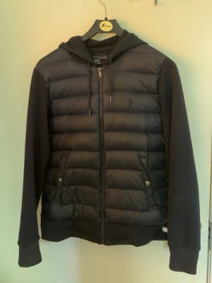 Mörkblå ralph lauren cardigan - Mörkblå ralph lauren jacka storlek S med huva och dragkedja framtill. Jackan har quiltad front, släta ärmar och två fickor med tryckknappar. Perfekt för kalla dagar och har en sportig, modern look.