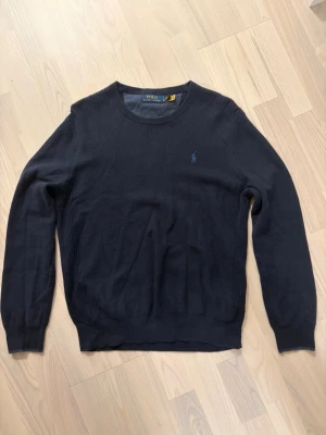 Mörkblå tröja från Polo Ralph Lauren - Mörkblå stickad tröja från Polo Ralph Lauren med rund halsringning och diskret broderad logga på bröstet. Tröjan har långa ärmar och ribbade muddar vid ärmslut och nederkant. Perfekt för en stilren och klassisk look.