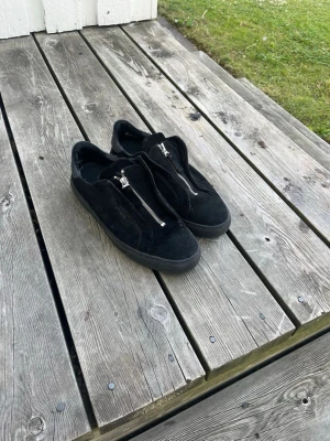 Axel arigato skor - Snygga svarta sneakers i mocka med silverfärgad dragkedja framtill istället för klassiska snören. Skorna har en låg profil och svart sula, vilket ger en clean och modern look. Perfekta för dig som gillar stilrena detaljer och enkel design.