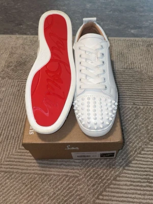 Vita sneakers med nitar och röd sula - Stilrena vita sneakers med coola nitar på tån och klassisk röd sula. Skorna har vita skosnören och är tillverkade i slätt läder. Perfekta för dig som vill sticka ut med en edgy och trendig look.