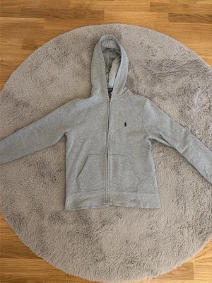 En knappt använd grå Ralph lauren zip hoodie med huva. - Säljer en stilren grå hoodie med dragkedja och huva. Tröjan har två fickor framtill och en liten broderad logga på bröstet. Perfekt för chill dagar eller när du vill ha en enkel och snygg look. Mjuk bomullsblandning för extra komfort.