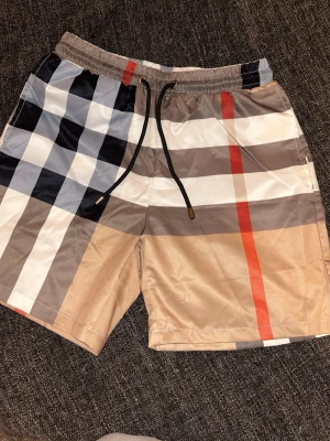 Rutiga badbyxor med snörning - Snygga badbyxor med klassiskt rutigt mönster i beige, svart, vitt och rött. De har elastisk midja med svart dragsko och en bakficka. Materialet är snabbtorkande och perfekt för stranden eller poolen. Passar dig som gillar stilrena och trendiga badkläder. Burrberry shorts sitter som S 