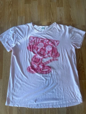 Vit Mickey Mouse t-shirt Disney - Säljer en vit t-shirt från Disney med ett stort rosa tryck av Mickey Mouse på framsidan. T-shirten har klassisk passform och korta ärmar. Perfekt för dig som gillar retro och tecknade motiv.